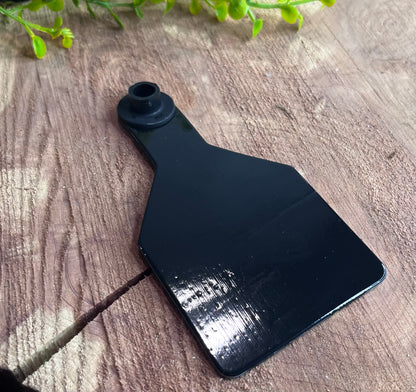 Black plastic medium cow tag on a wooden surface with greenery in the corner