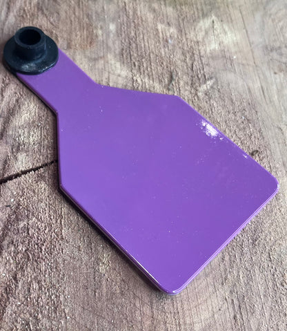 Purple plastic medium cow tag on a wooden surface with greenery in the corner