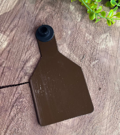 Brown plastic medium cow tag on a wooden surface with greenery in the corner