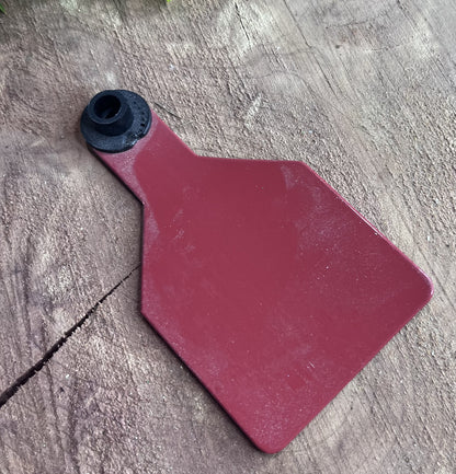 Red plastic medium cow tag on a wooden surface with greenery in the corner