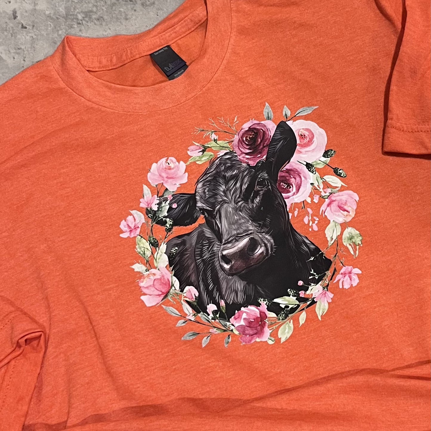 Orange t-shirt with a black cow surrounded by pink flowers on a concrete surface