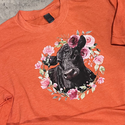 Orange t-shirt with a black cow surrounded by pink flowers on a concrete surface